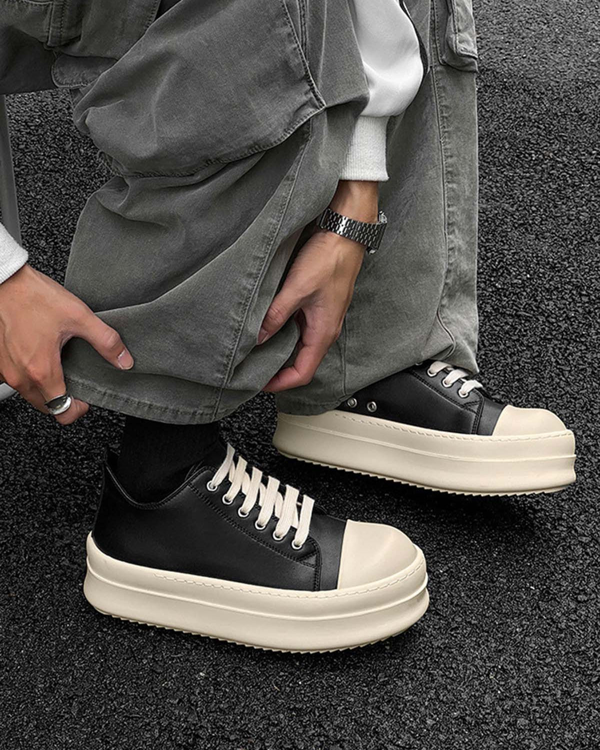 Person crouching, wearing Chunky Low-Top Streetwear Sneakers with black uppers, white laces, and thick off-white soles, paired with loose grey pants and accessories.