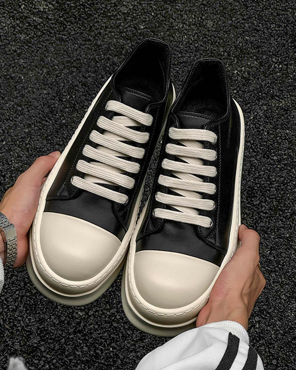 Person holding Chunky Low-Top Streetwear Sneakers with black uppers, off-white thick rubber soles, and toe caps, showcasing a German-trainer inspired design over a textured dark floor.
