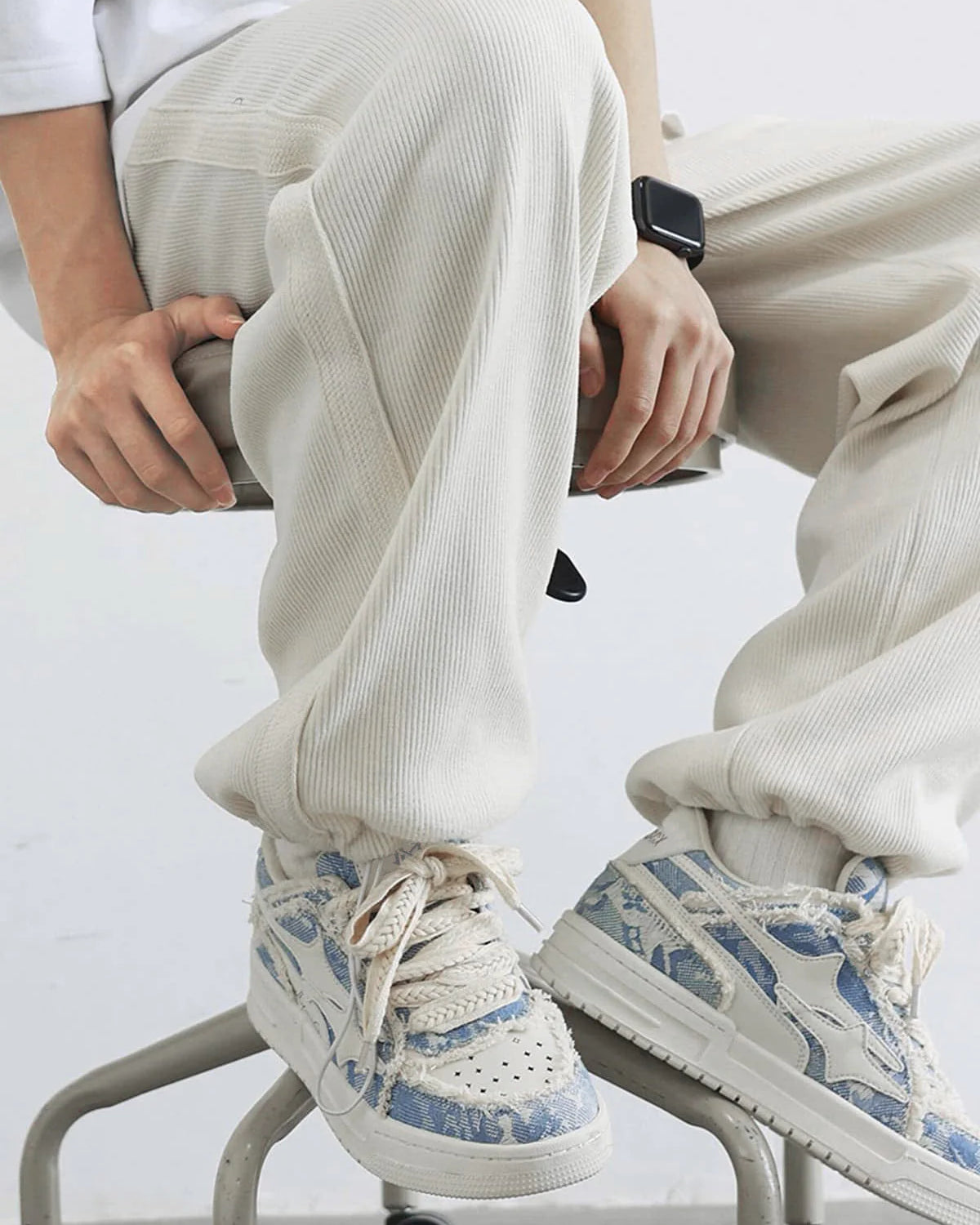 Distressed Denim Low-Top Street Sneakers with frayed denim-print, chunky soles, and braided laces, showcased with rib-knit joggers on a rolling stool.