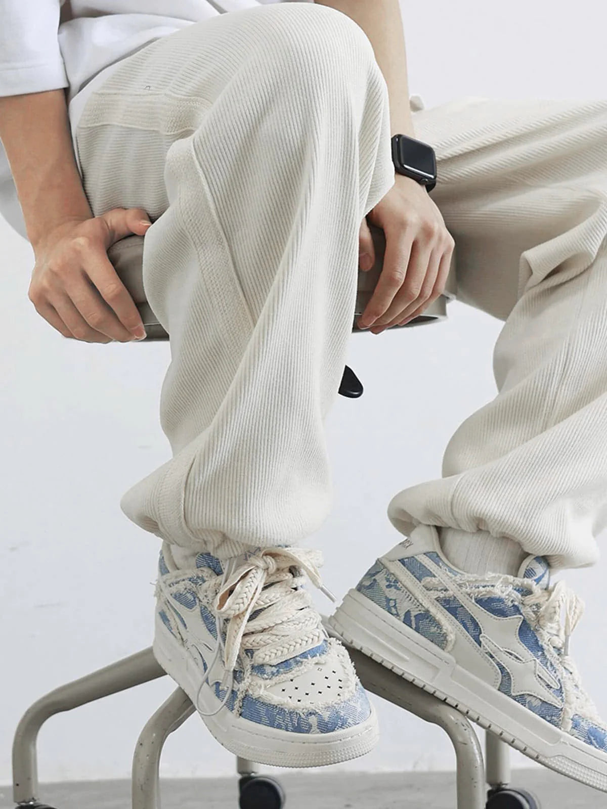 Distressed Denim Low-Top Street Sneakers with frayed denim-print, chunky soles, and braided laces, showcased with rib-knit joggers on a rolling stool.