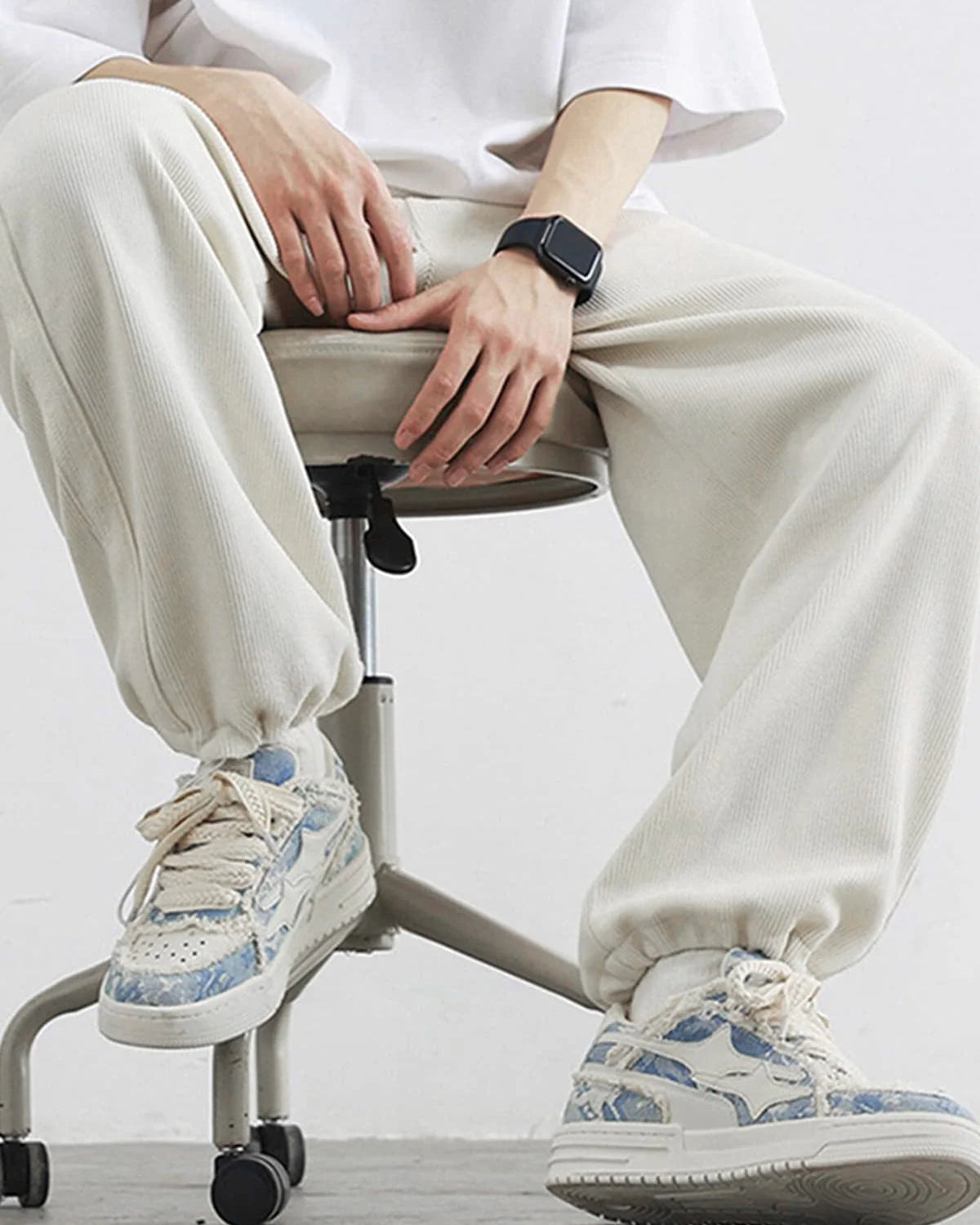 Seated person wearing Distressed Denim Low-Top Street Sneakers with chunky design and pale blue accents, paired with light-cream jogger-style pants and a smartwatch.