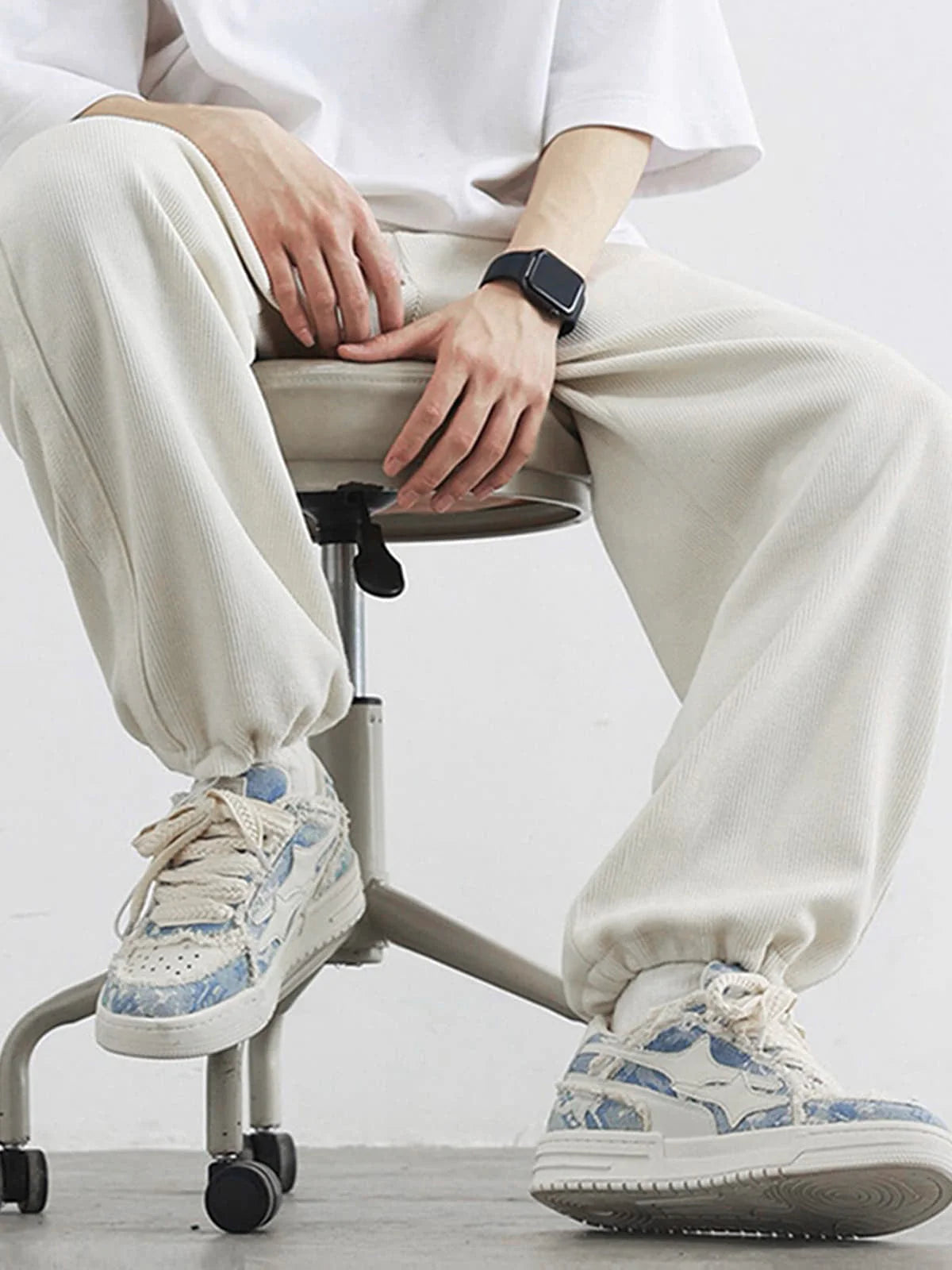 Seated person wearing Distressed Denim Low-Top Street Sneakers with chunky design and pale blue accents, paired with light-cream jogger-style pants and a smartwatch.