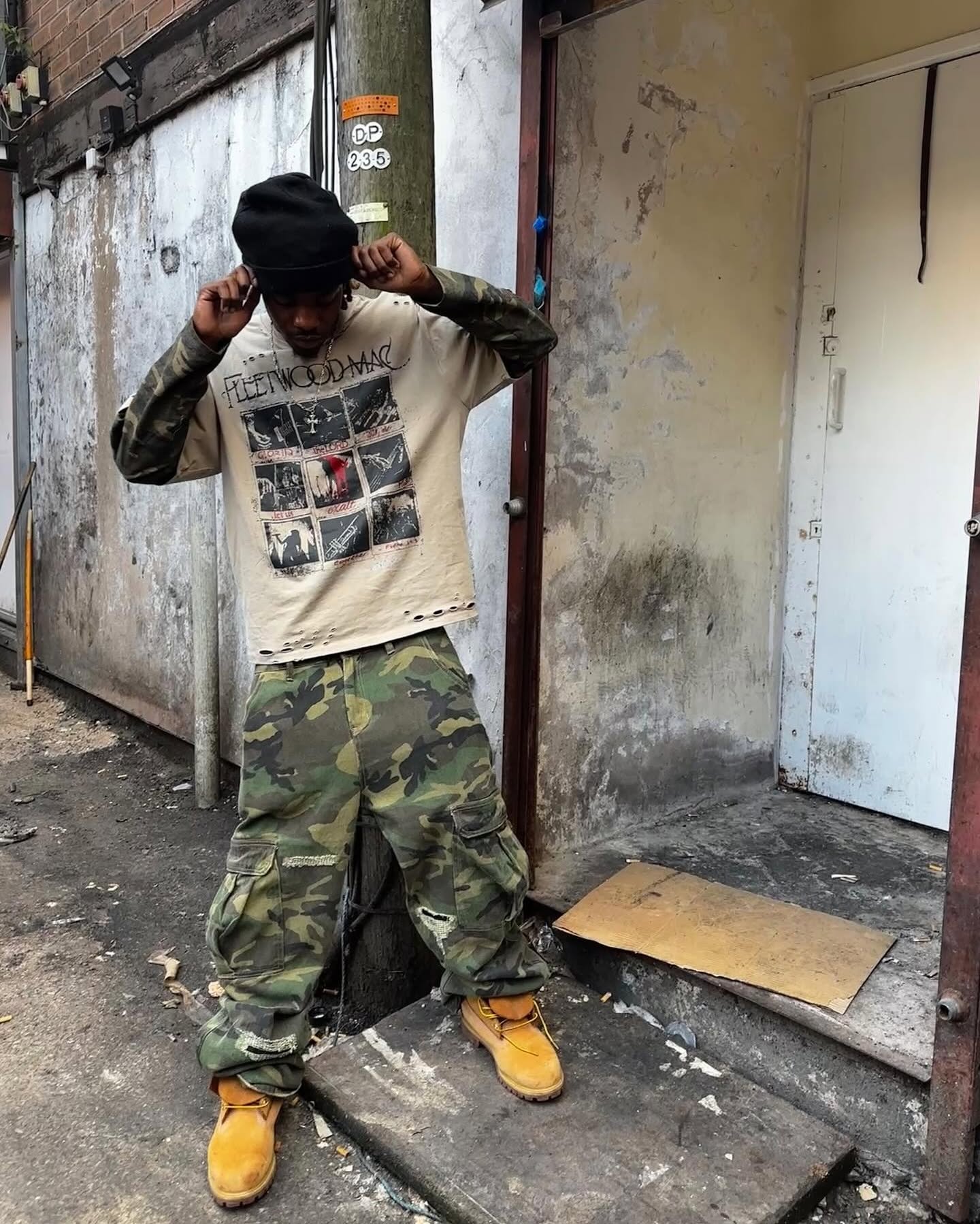 Young man wearing a Distressed Grunge Layered T-Shirt with camo sleeves, standing urban style in an alley, adjusting his beanie.