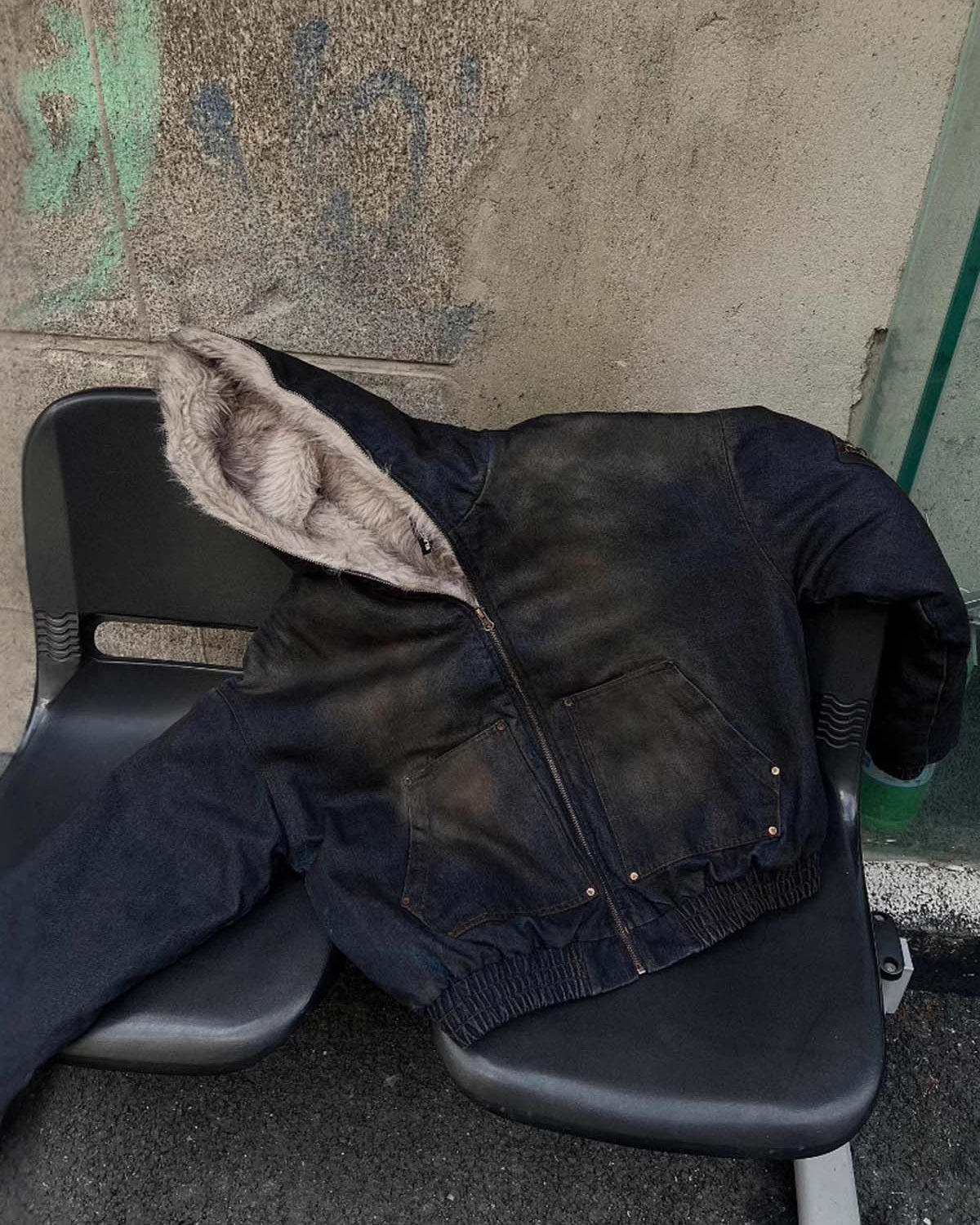 Distressed Washed Faux Fur Winter Jacket with a hood, fleece-lined interior, front pockets, and elasticized hem, displayed on plastic seats against a graffiti-marked wall.