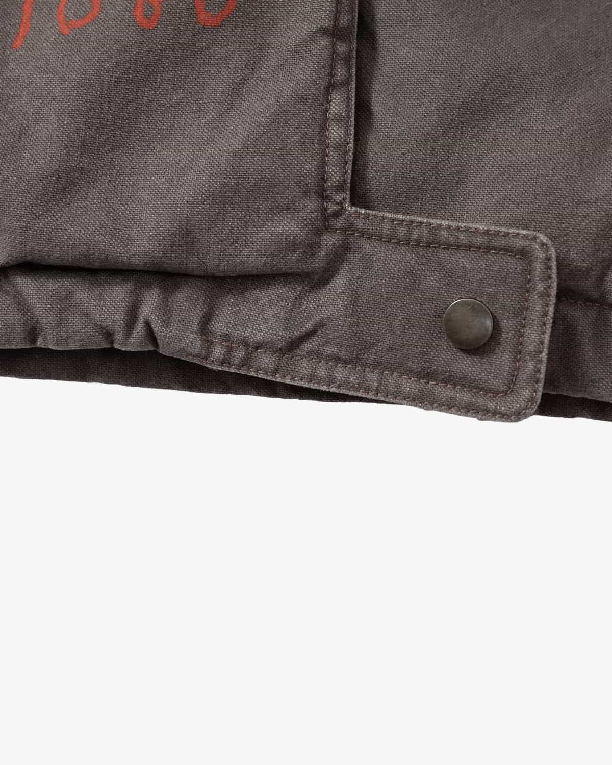 Close-up of Graphic Graffiti High Neck Street Jacket hem, showing folded canvas fabric, reinforced stitching, and adjustable snap-closure tab with red branding detail.