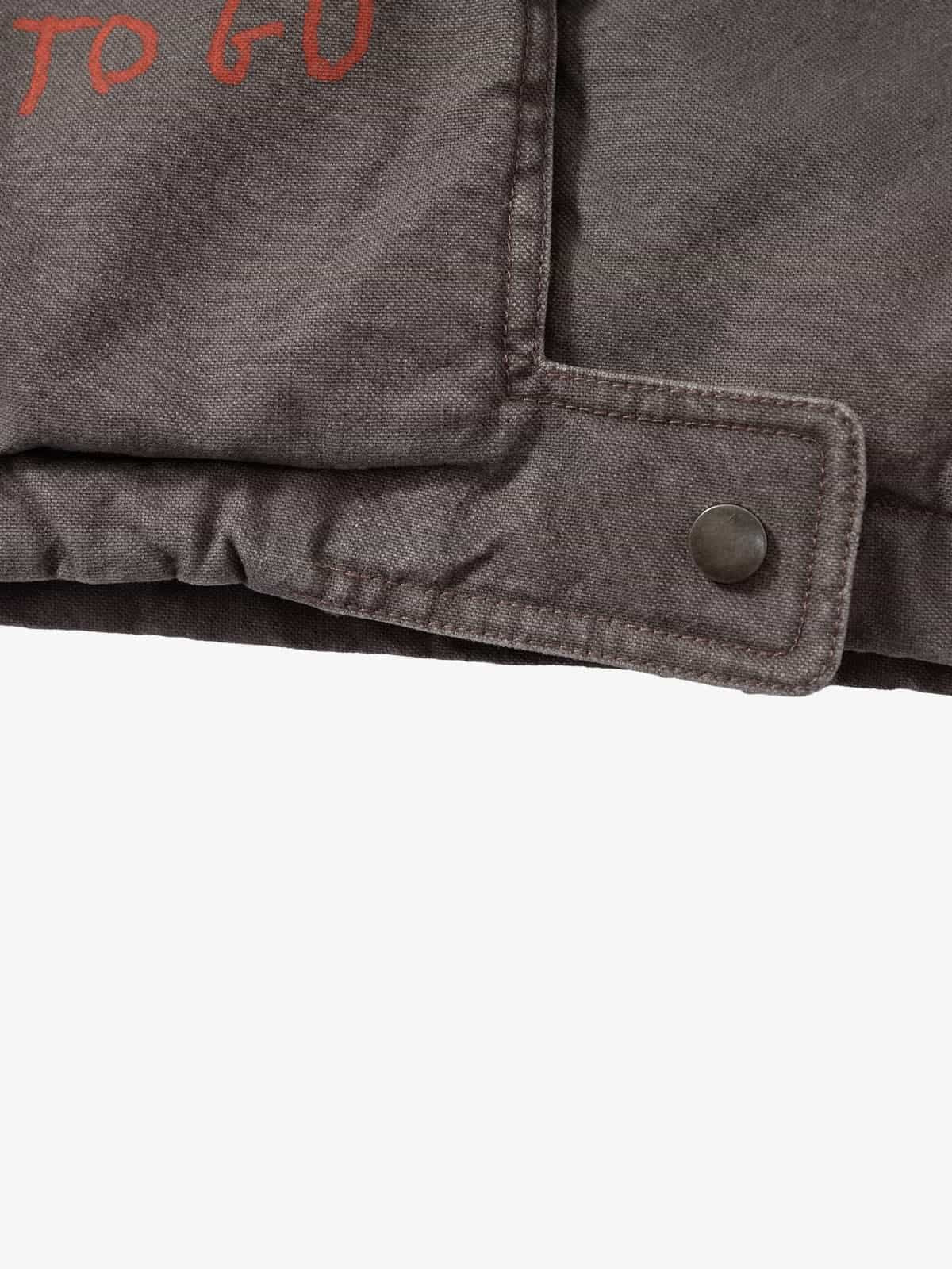 Close-up of Graphic Graffiti High Neck Street Jacket hem, showing folded canvas fabric, reinforced stitching, and adjustable snap-closure tab with red branding detail.