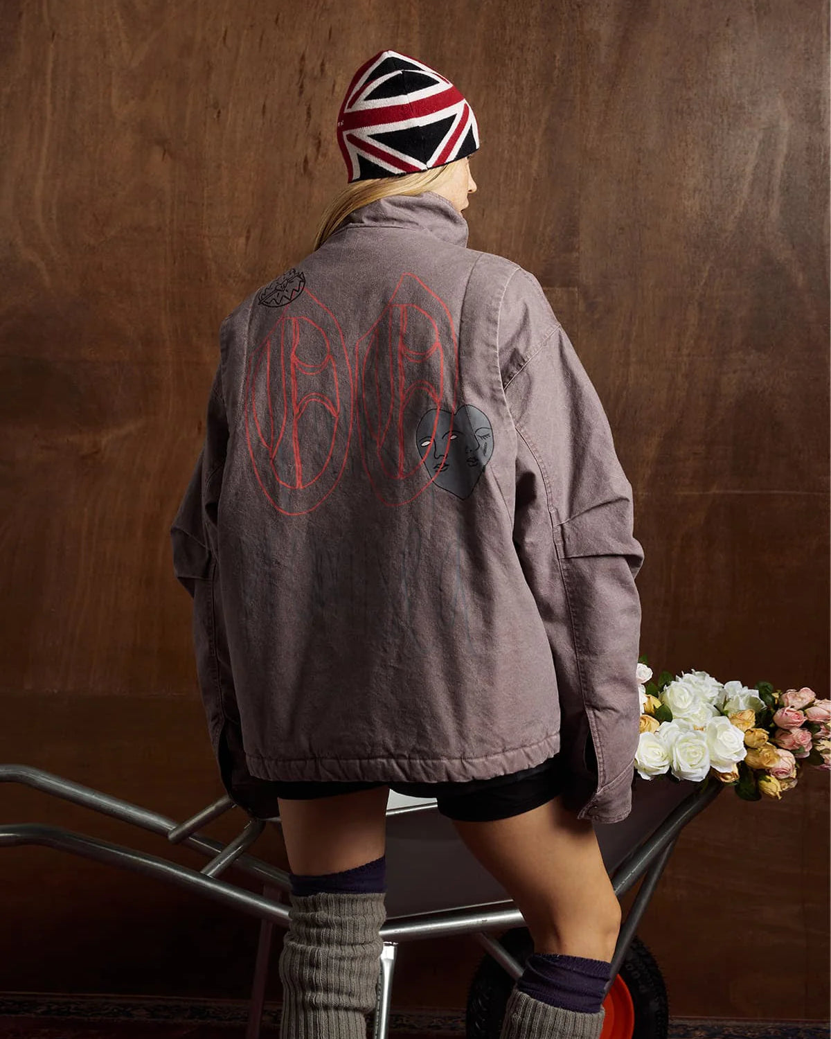 Person wearing the Graphic Graffiti High Neck Street Jacket, showcasing bold graffiti designs and a high-neck silhouette, while pushing a wheelbarrow filled with roses indoors.
