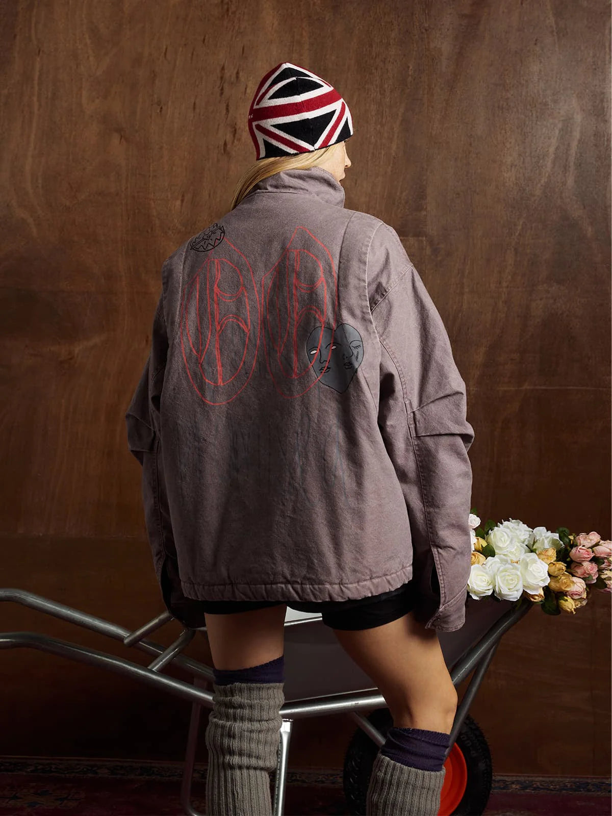 Person wearing the Graphic Graffiti High Neck Street Jacket, showcasing bold graffiti designs and a high-neck silhouette, while pushing a wheelbarrow filled with roses indoors.