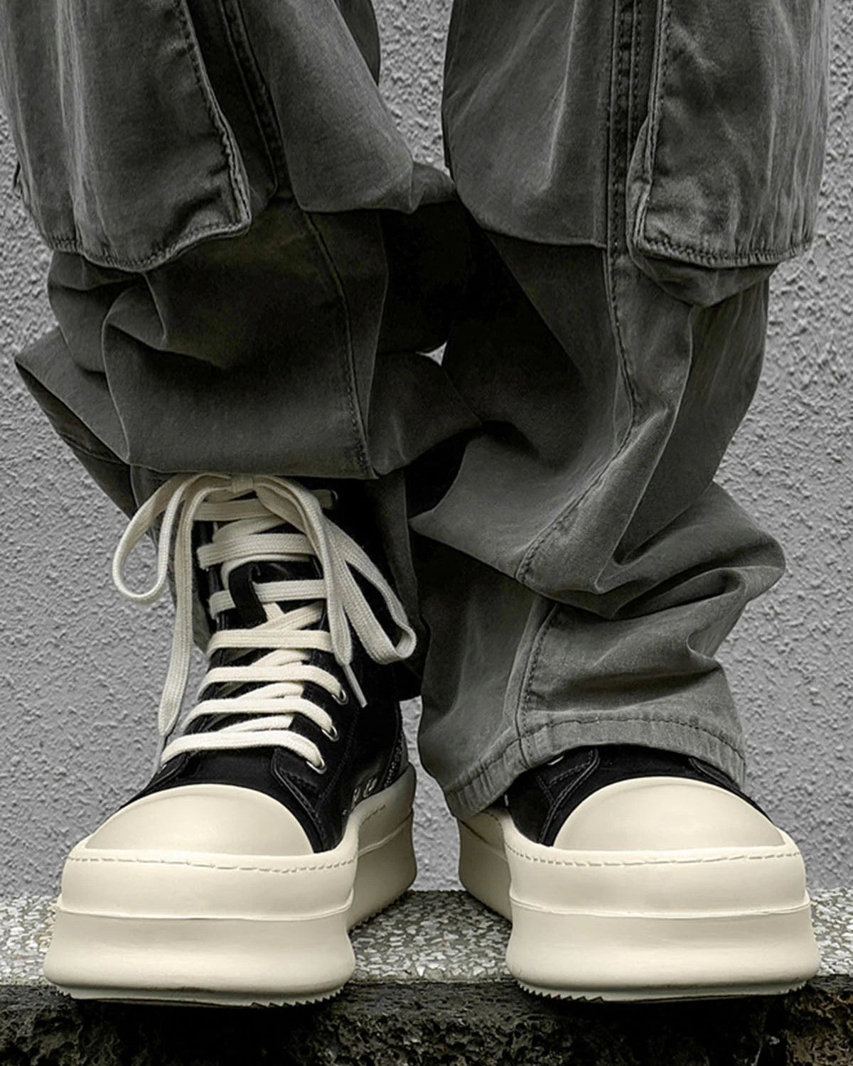 Person wears High-Top Platform Streetwear Sneakers with oversized cream platform soles, loose grayish-green pants draping over them, standing on a stone surface.