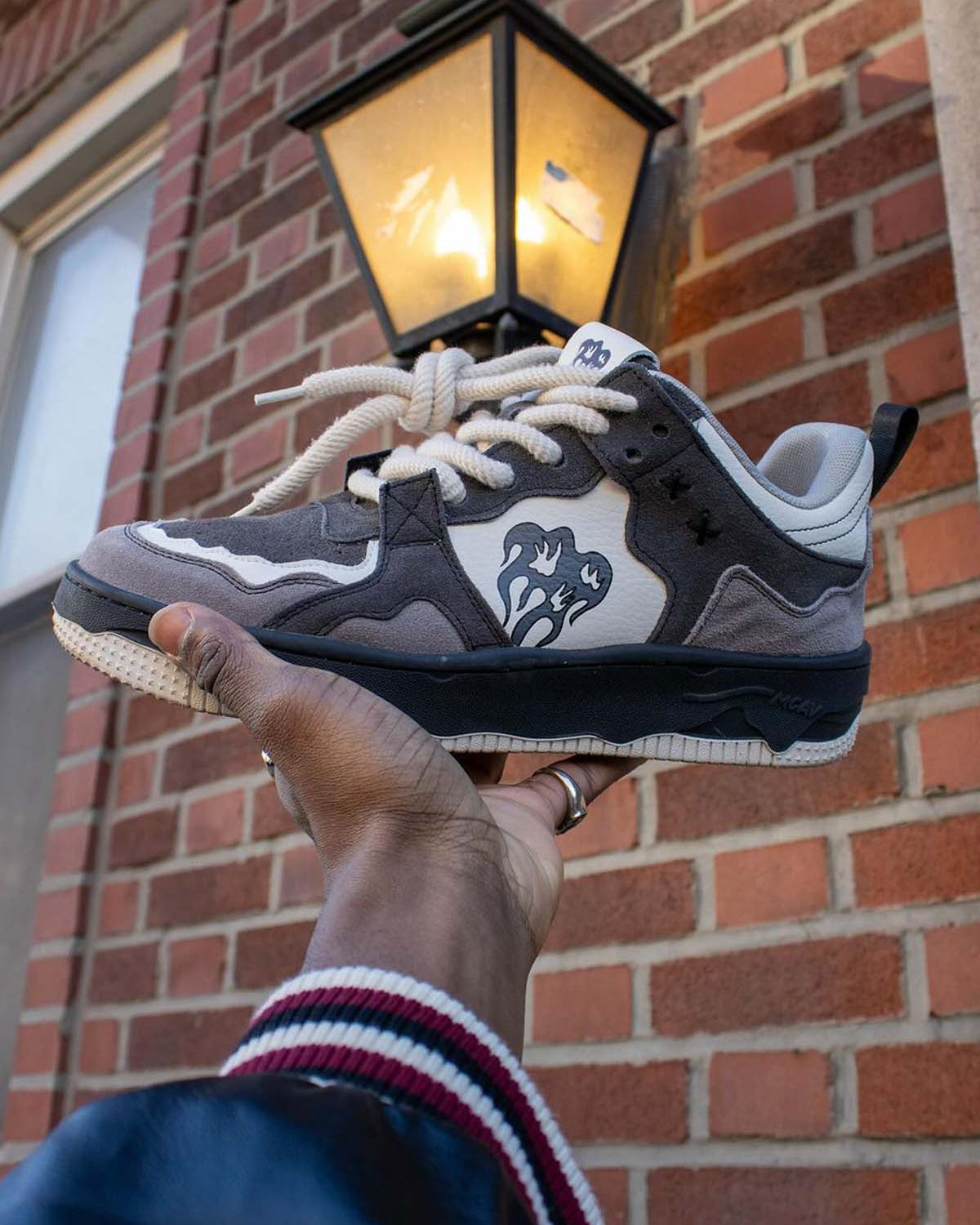 Person holding Vintage Chunky Graphic Skate Shoes, showcasing their grey, black, and off-white panels with chunky laces and a dripping-heart logo against a brick wall.