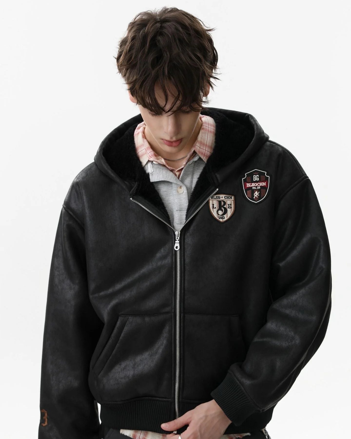 Young man in a Vintage Hooded Leather-Look Street Jacket with shearling-lined hood, vintage patches, layered over a plaid shirt, showcasing street style.