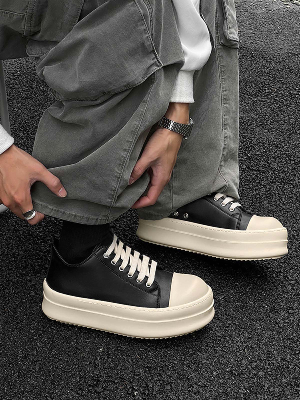 Person crouching, wearing Chunky Low-Top Streetwear Sneakers with black uppers, white laces, and thick off-white soles, paired with loose grey pants and accessories.