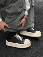 Person crouching, wearing Chunky Low-Top Streetwear Sneakers with black uppers, white laces, and thick off-white soles, paired with loose grey pants and accessories.