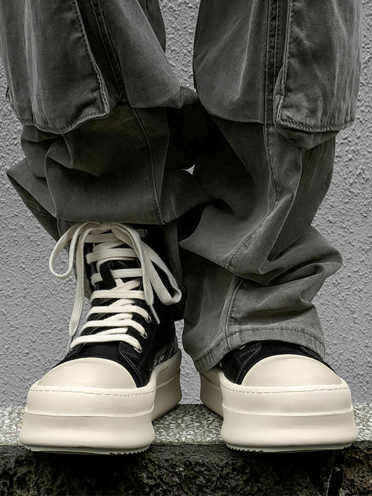 Person wears High-Top Platform Streetwear Sneakers with oversized cream platform soles, loose grayish-green pants draping over them, standing on a stone surface.