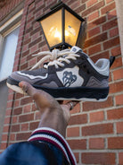 Person holding Vintage Chunky Graphic Skate Shoes, showcasing their grey, black, and off-white panels with chunky laces and a dripping-heart logo against a brick wall.